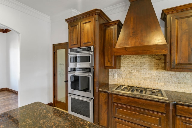 kitchen with arched walkways, crown molding, custom exhaust hood, stainless steel appliances, and decorative backsplash