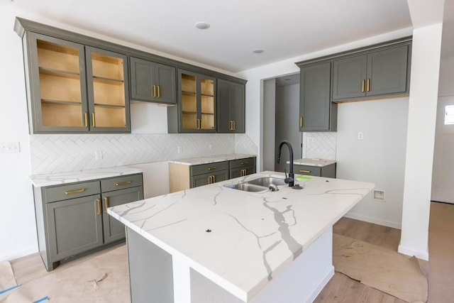 kitchen with a kitchen island with sink, sink, decorative backsplash, light hardwood / wood-style floors, and light stone counters