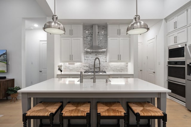 kitchen featuring appliances with stainless steel finishes, an island with sink, hanging light fixtures, and wall chimney range hood