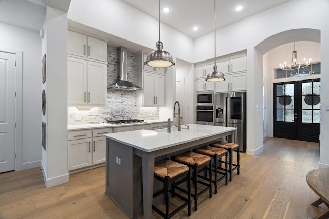 kitchen with appliances with stainless steel finishes, light wood-type flooring, wall chimney exhaust hood, a center island with sink, and white cabinetry