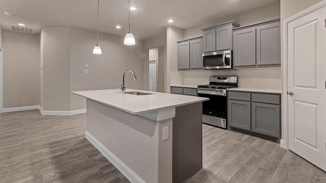 kitchen featuring appliances with stainless steel finishes, light wood-type flooring, sink, hanging light fixtures, and an island with sink