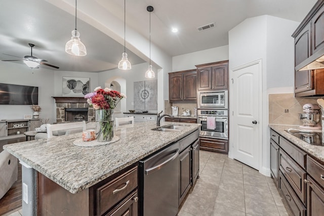 kitchen with sink, hanging light fixtures, appliances with stainless steel finishes, a fireplace, and a kitchen island with sink