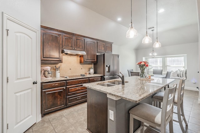 kitchen featuring sink, a kitchen island with sink, light stone counters, dark brown cabinetry, and stainless steel refrigerator with ice dispenser