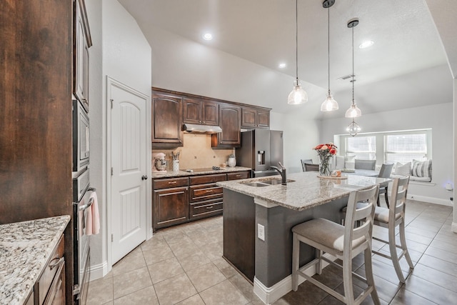 kitchen with dark brown cabinetry, hanging light fixtures, a center island with sink, appliances with stainless steel finishes, and light stone countertops