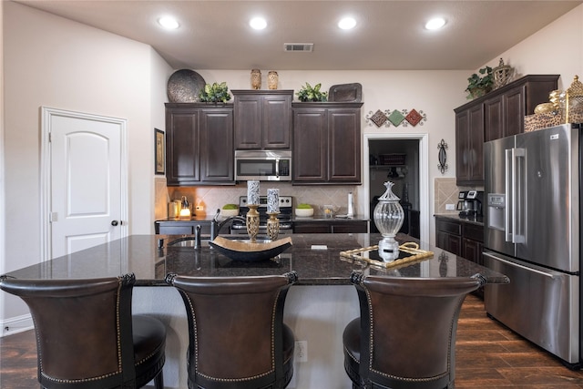 kitchen with a center island with sink, dark hardwood / wood-style floors, stainless steel appliances, and dark stone counters