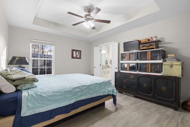 bedroom with light hardwood / wood-style floors, connected bathroom, ceiling fan, and a tray ceiling