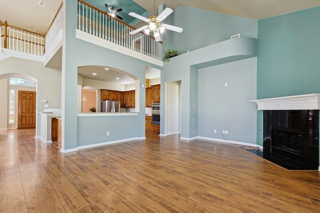 unfurnished living room featuring a high ceiling, a tiled fireplace, ceiling fan, and hardwood / wood-style floors