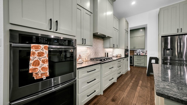 kitchen featuring dark wood-type flooring, premium range hood, dark stone counters, decorative backsplash, and appliances with stainless steel finishes