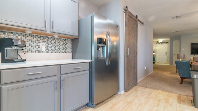 kitchen with a barn door, gray cabinets, decorative backsplash, light wood-type flooring, and stainless steel fridge with ice dispenser