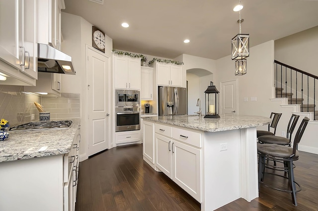 kitchen featuring dark wood-type flooring, a center island with sink, appliances with stainless steel finishes, decorative light fixtures, and white cabinetry