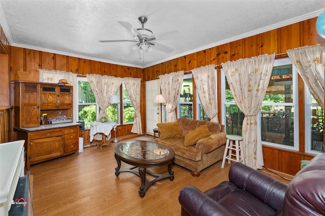 living room featuring wood walls, light wood-type flooring, ornamental molding, ceiling fan, and a textured ceiling