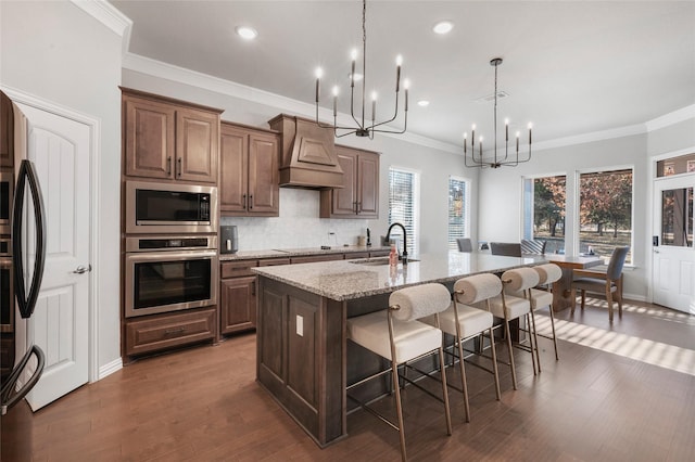 kitchen with dark wood-type flooring, stainless steel appliances, decorative light fixtures, a center island with sink, and custom exhaust hood