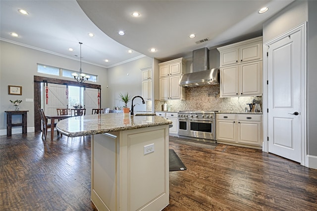 kitchen featuring wall chimney range hood, light stone counters, decorative light fixtures, range with two ovens, and a center island with sink