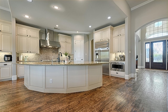 kitchen featuring dark wood-type flooring, a center island with sink, built in appliances, wall chimney exhaust hood, and ornamental molding