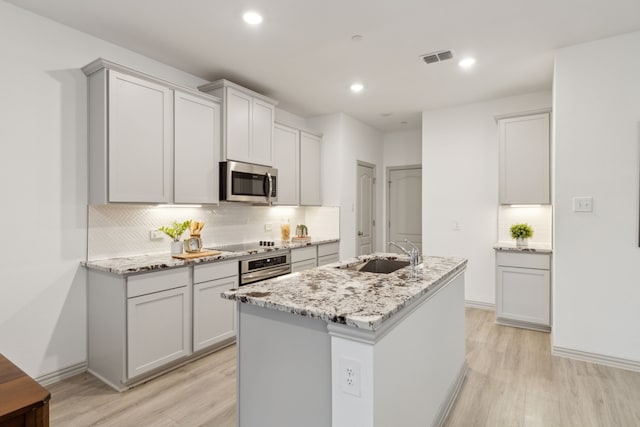 kitchen with light stone counters, stainless steel appliances, a kitchen island with sink, sink, and light hardwood / wood-style flooring