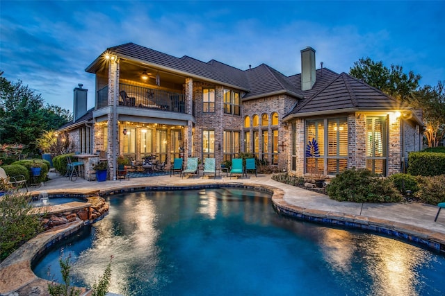 back house at dusk featuring ceiling fan, a balcony, a swimming pool with hot tub, and a patio area