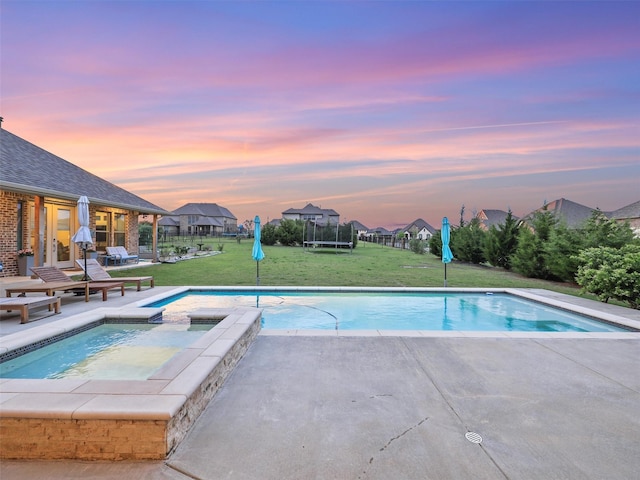 pool at dusk featuring an in ground hot tub, a trampoline, a patio area, and a lawn