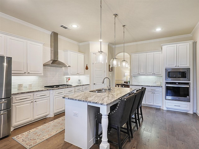 kitchen with sink, wall chimney range hood, a kitchen island with sink, white cabinets, and appliances with stainless steel finishes