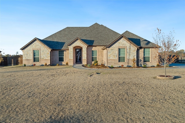 french country home with stone siding, brick siding, roof with shingles, and fence