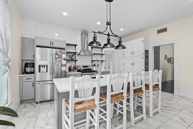 kitchen featuring gray cabinetry, wall chimney range hood, backsplash, a kitchen island with sink, and appliances with stainless steel finishes