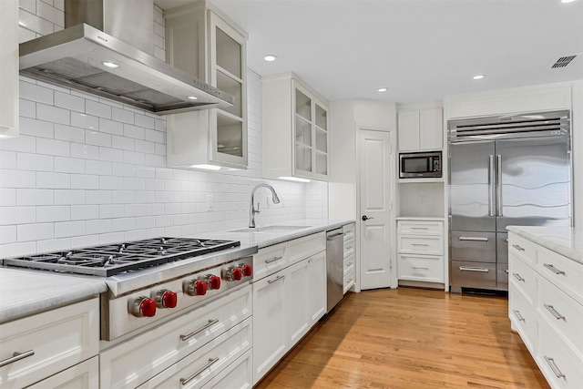 kitchen with sink, white cabinetry, built in appliances, light stone countertops, and wall chimney exhaust hood