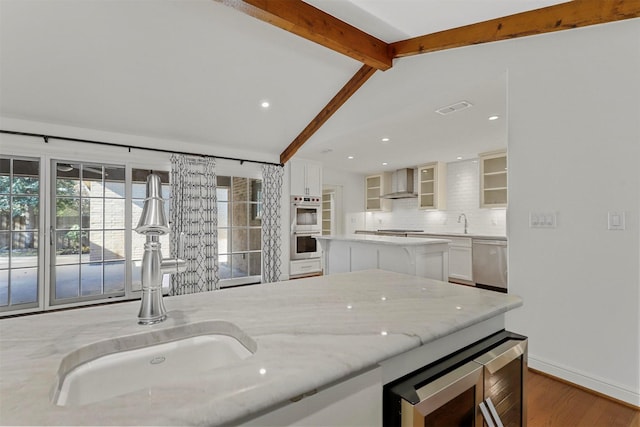 kitchen with light stone counters, lofted ceiling with beams, appliances with stainless steel finishes, wall chimney range hood, and white cabinets