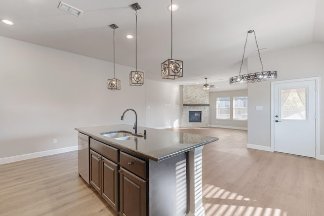 kitchen with a stone fireplace, an island with sink, sink, hanging light fixtures, and stainless steel dishwasher