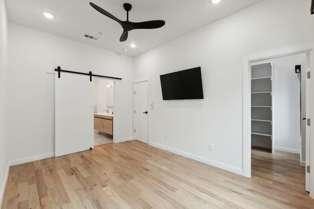 unfurnished bedroom featuring connected bathroom, a barn door, light wood-type flooring, ceiling fan, and a spacious closet