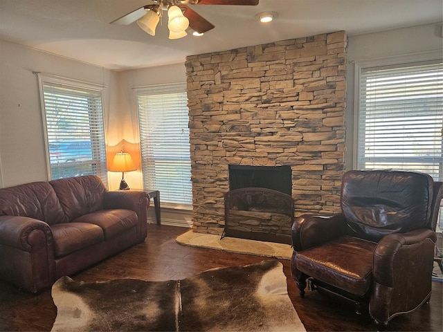 living room featuring dark wood-type flooring, a stone fireplace, ceiling fan, and a healthy amount of sunlight