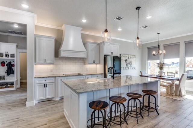 kitchen with white cabinets, custom range hood, stainless steel refrigerator with ice dispenser, and an island with sink