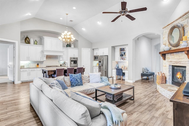 living room with a stone fireplace, ceiling fan with notable chandelier, high vaulted ceiling, and light hardwood / wood-style flooring