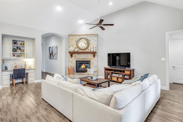 living room with ceiling fan, high vaulted ceiling, built in desk, light hardwood / wood-style floors, and a stone fireplace