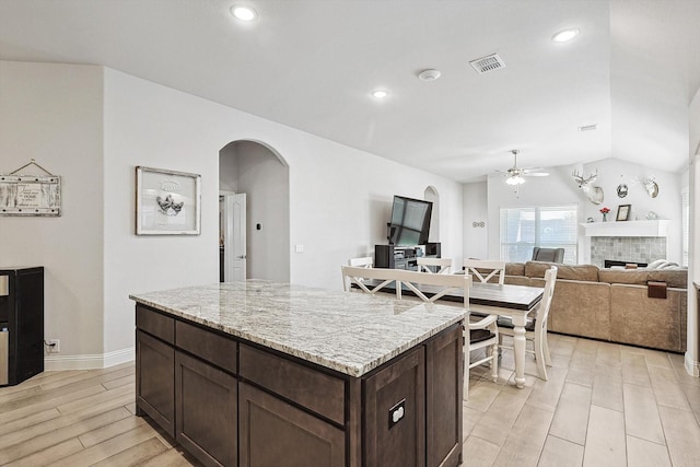 kitchen with dark brown cabinetry, lofted ceiling, light stone counters, a tile fireplace, and ceiling fan