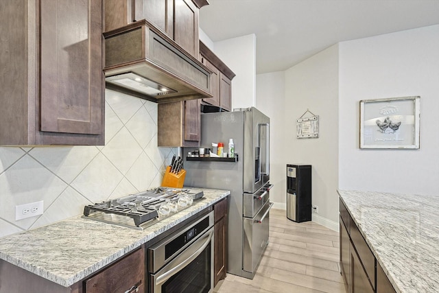 kitchen featuring decorative backsplash, dark brown cabinetry, light stone counters, stainless steel appliances, and custom range hood