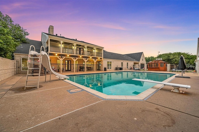 pool at dusk featuring a diving board, a patio area, and a water slide