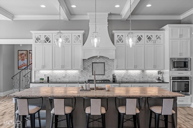 kitchen with beamed ceiling, white cabinetry, stainless steel appliances, and a kitchen island with sink