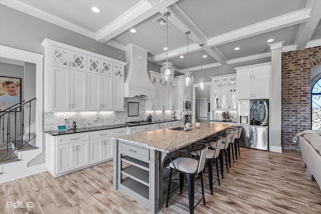 kitchen featuring sink, beamed ceiling, premium range hood, an island with sink, and light hardwood / wood-style floors