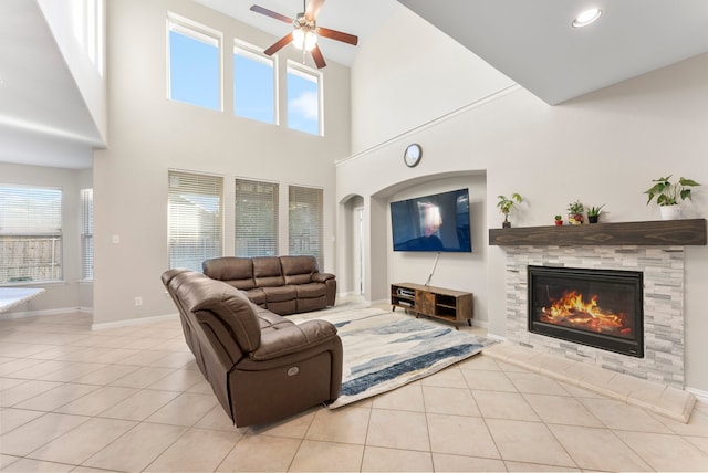 living room with ceiling fan, a stone fireplace, light tile patterned floors, and a towering ceiling