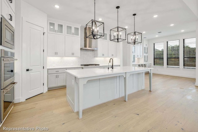 kitchen with hanging light fixtures, wall chimney range hood, a kitchen island with sink, white cabinets, and light wood-type flooring