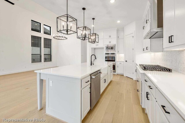 kitchen with appliances with stainless steel finishes, wall chimney range hood, light hardwood / wood-style flooring, hanging light fixtures, and an island with sink