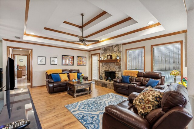 living room with ceiling fan, a raised ceiling, a stone fireplace, light wood-type flooring, and ornamental molding