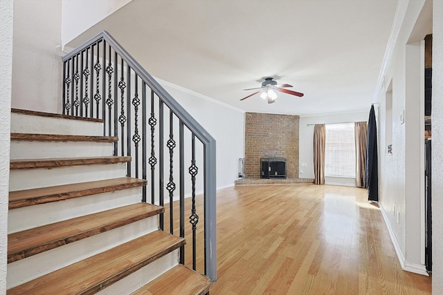 staircase with crown molding, ceiling fan, wood-type flooring, and a brick fireplace