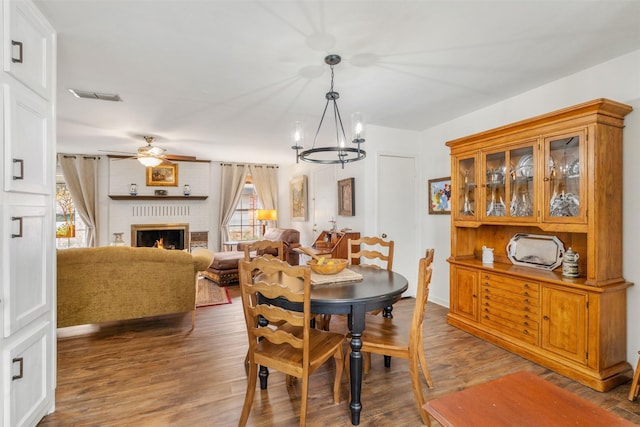 dining space with ceiling fan with notable chandelier, a brick fireplace, a wealth of natural light, and dark wood-type flooring