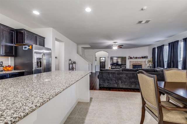 kitchen featuring light stone countertops, light wood-type flooring, ceiling fan, a fireplace, and stainless steel fridge with ice dispenser