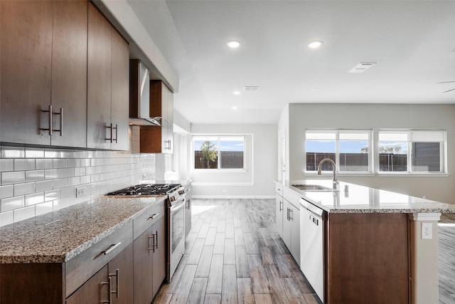 kitchen featuring sink, wall chimney exhaust hood, light stone counters, white dishwasher, and stainless steel stove