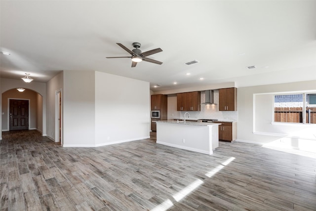 kitchen featuring ceiling fan, wall chimney exhaust hood, light hardwood / wood-style flooring, backsplash, and a kitchen island with sink