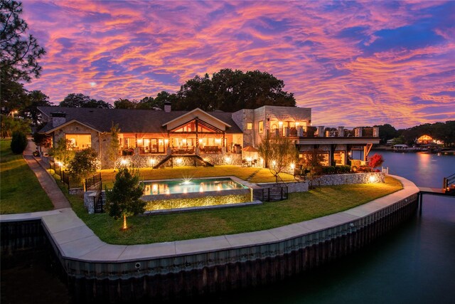 back house at dusk featuring a water view, a balcony, and a lawn