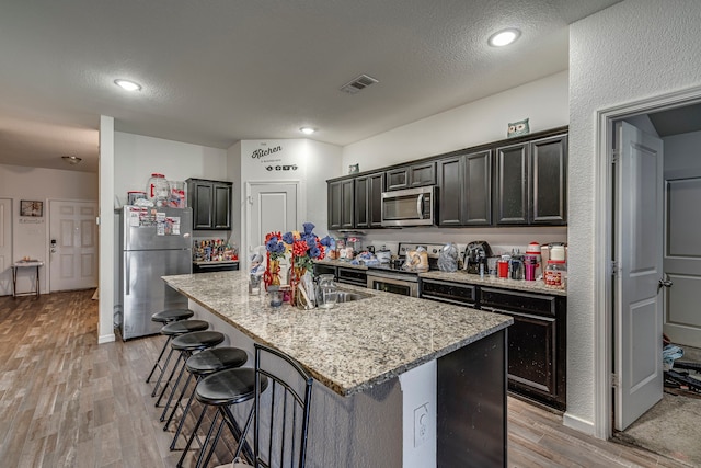 kitchen with appliances with stainless steel finishes, a kitchen island with sink, light hardwood / wood-style flooring, light stone counters, and sink