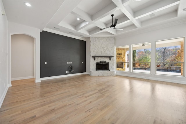 unfurnished living room featuring arched walkways, coffered ceiling, ceiling fan, a stone fireplace, and light wood-style floors