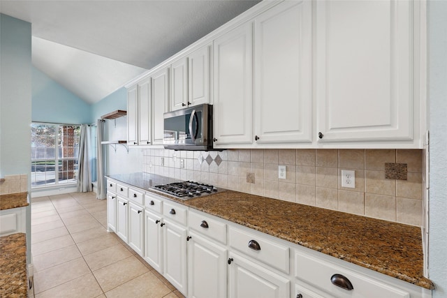 kitchen with appliances with stainless steel finishes, vaulted ceiling, light tile patterned floors, dark stone countertops, and white cabinets